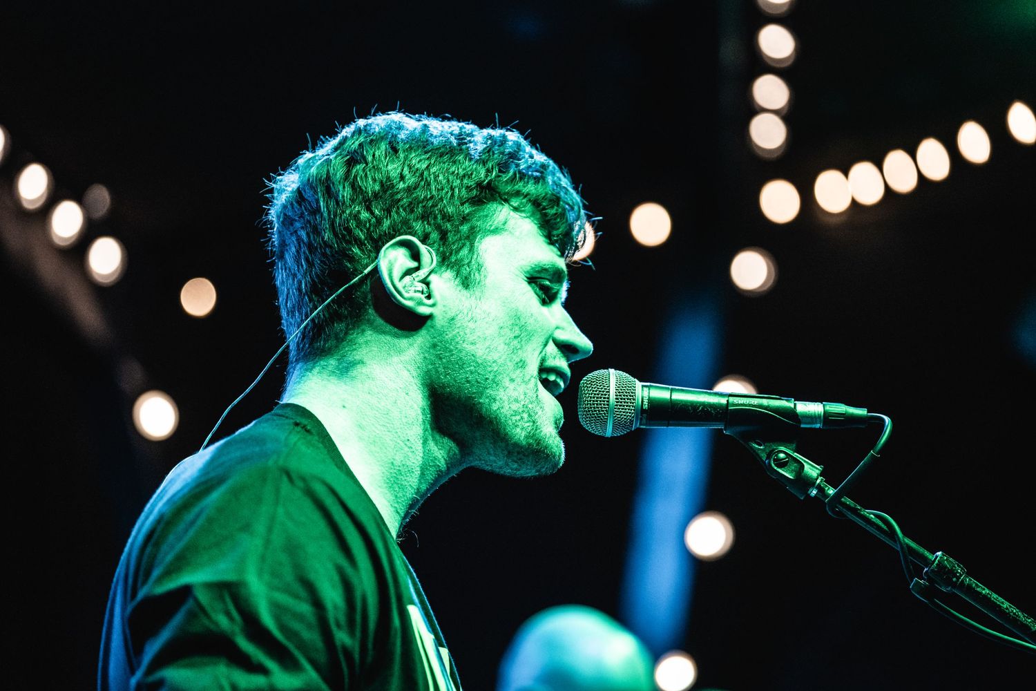 Performer singing into microphone on stage with green lighting and bokeh effects in background.