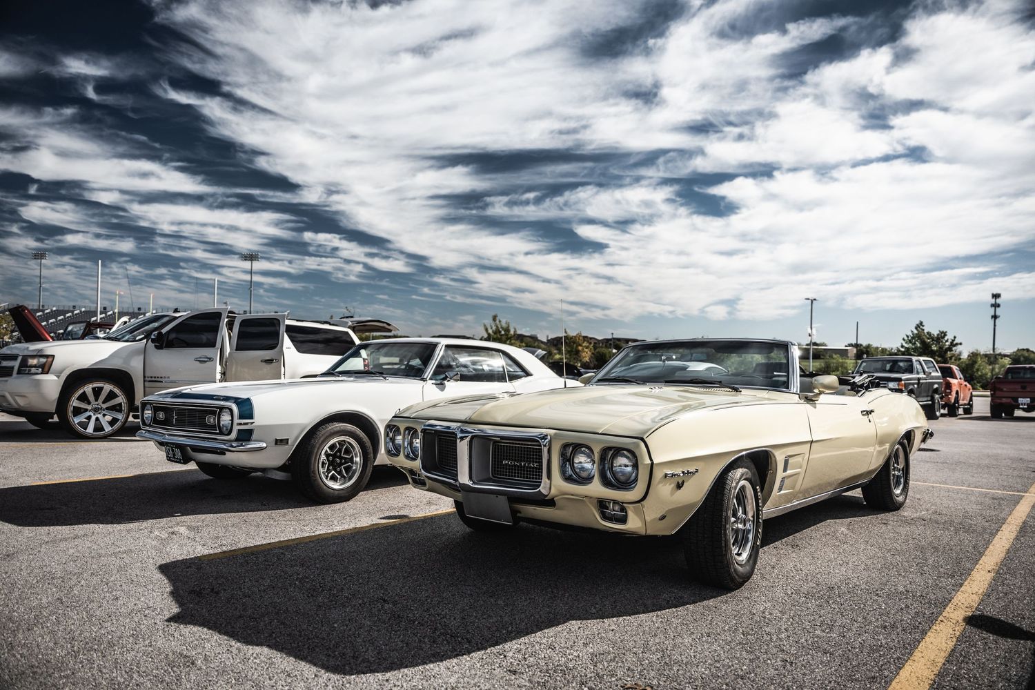 Classic Pontiac Firebird and vintage cars displayed at outdoor car show under dramatic cloudy sky.