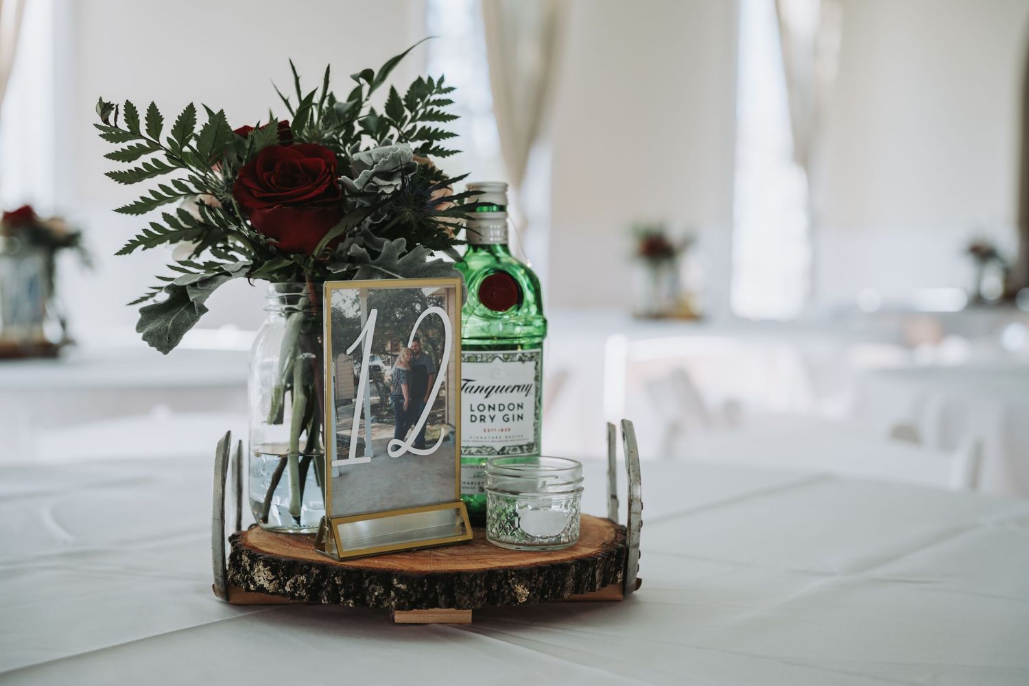 Rustic wedding centerpiece with wood slice, mason jars, and greenery on white tablecloth.