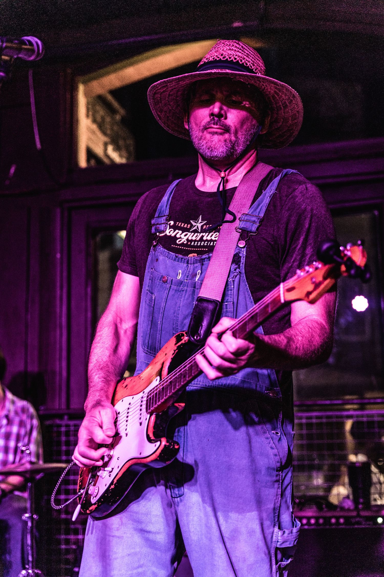 Musician in overalls and hat plays electric guitar under purple stage lighting.