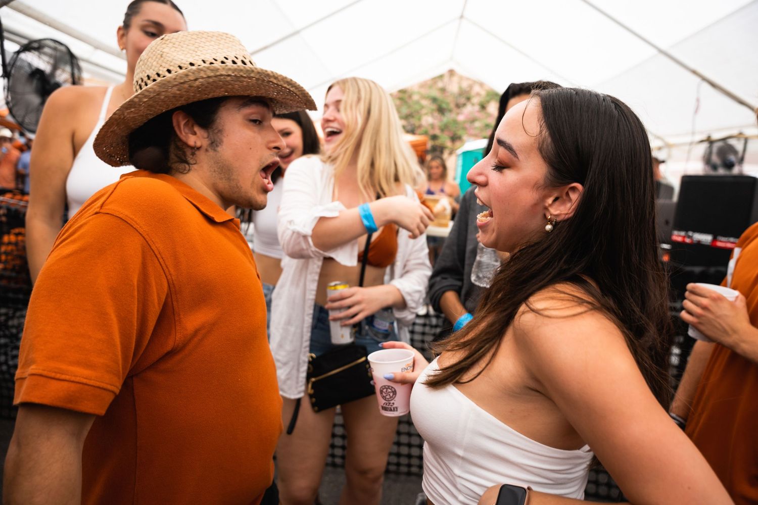 People enjoying drinks and socializing under a tent at an outdoor event wearing casual summer attire.