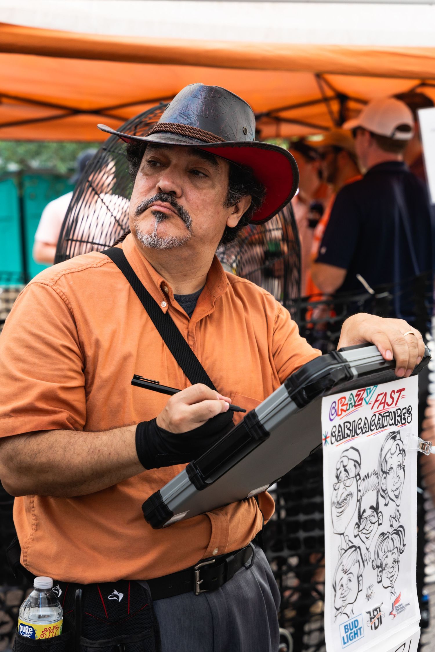 Caricature artist in orange shirt and cowboy hat drawing at outdoor event with sketch pad.
