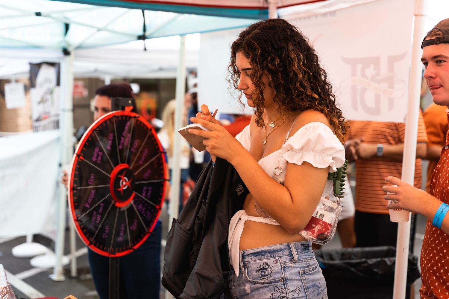 Young woman in white crop top checks phone at outdoor event with game wheel in background.