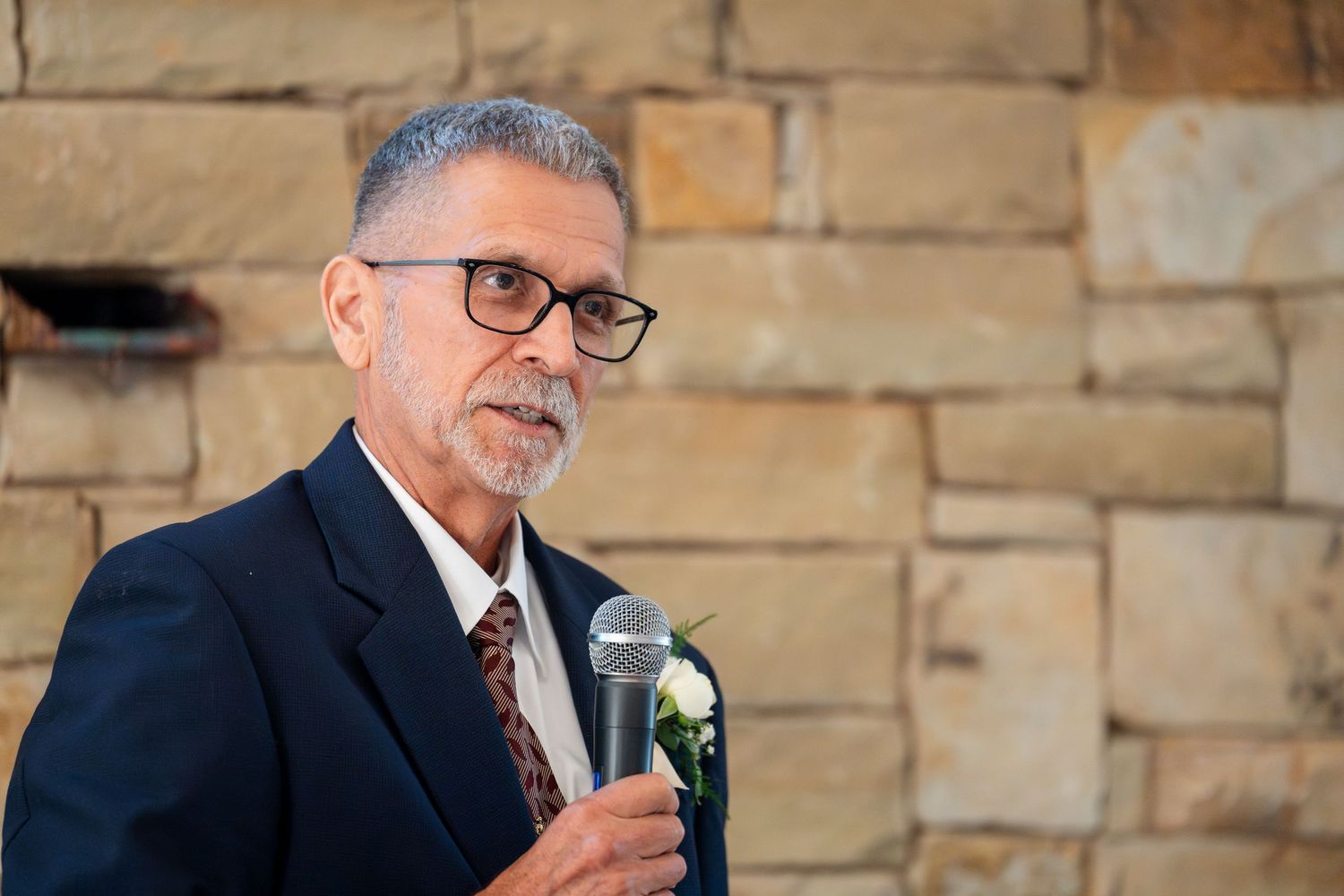 A speaker in glasses and dark suit gestures while giving a speech at a formal event.