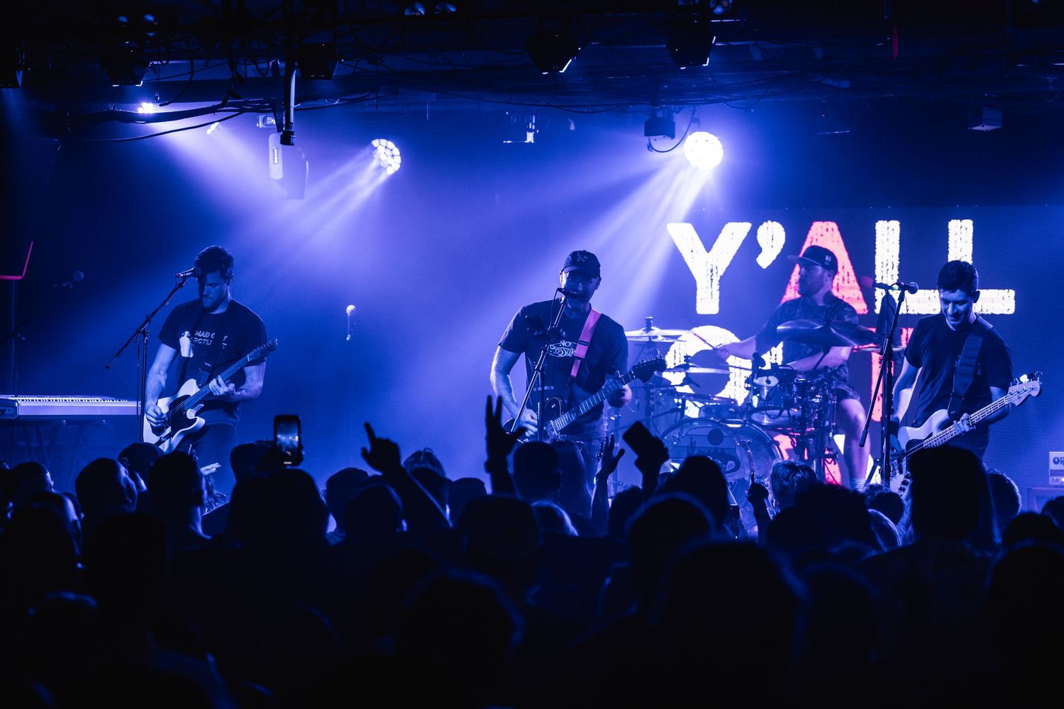 A rock band performs on stage with blue lighting and silhouetted crowd in foreground during a live concert.