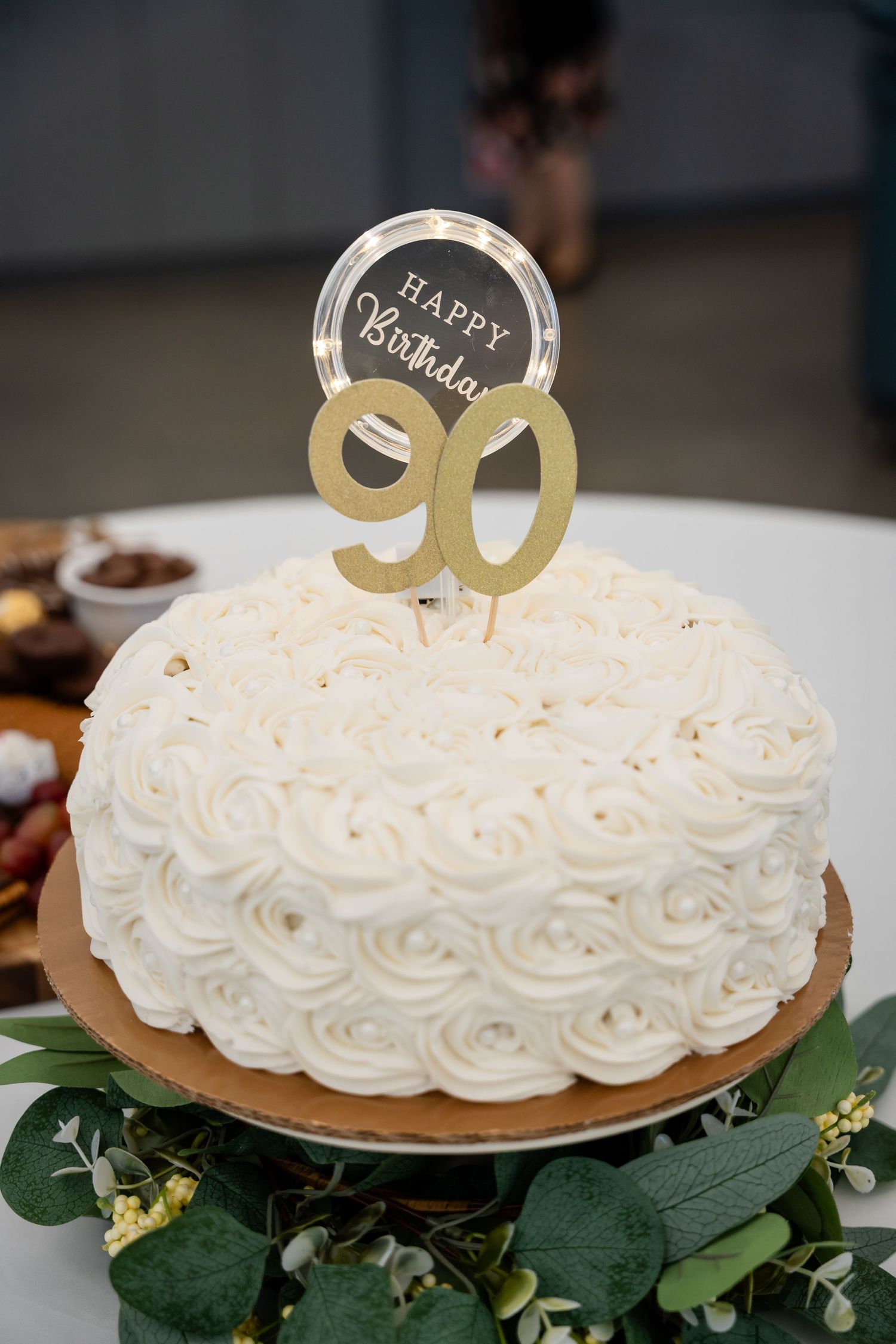 White rosette birthday cake with gold number 90 topper and greenery decorations.