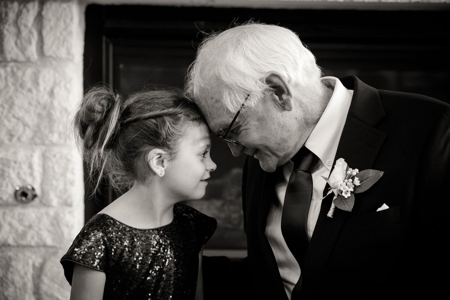 An elderly man in a suit leans down tenderly toward a young girl wearing a sparkly black dress in an intimate black and white moment.