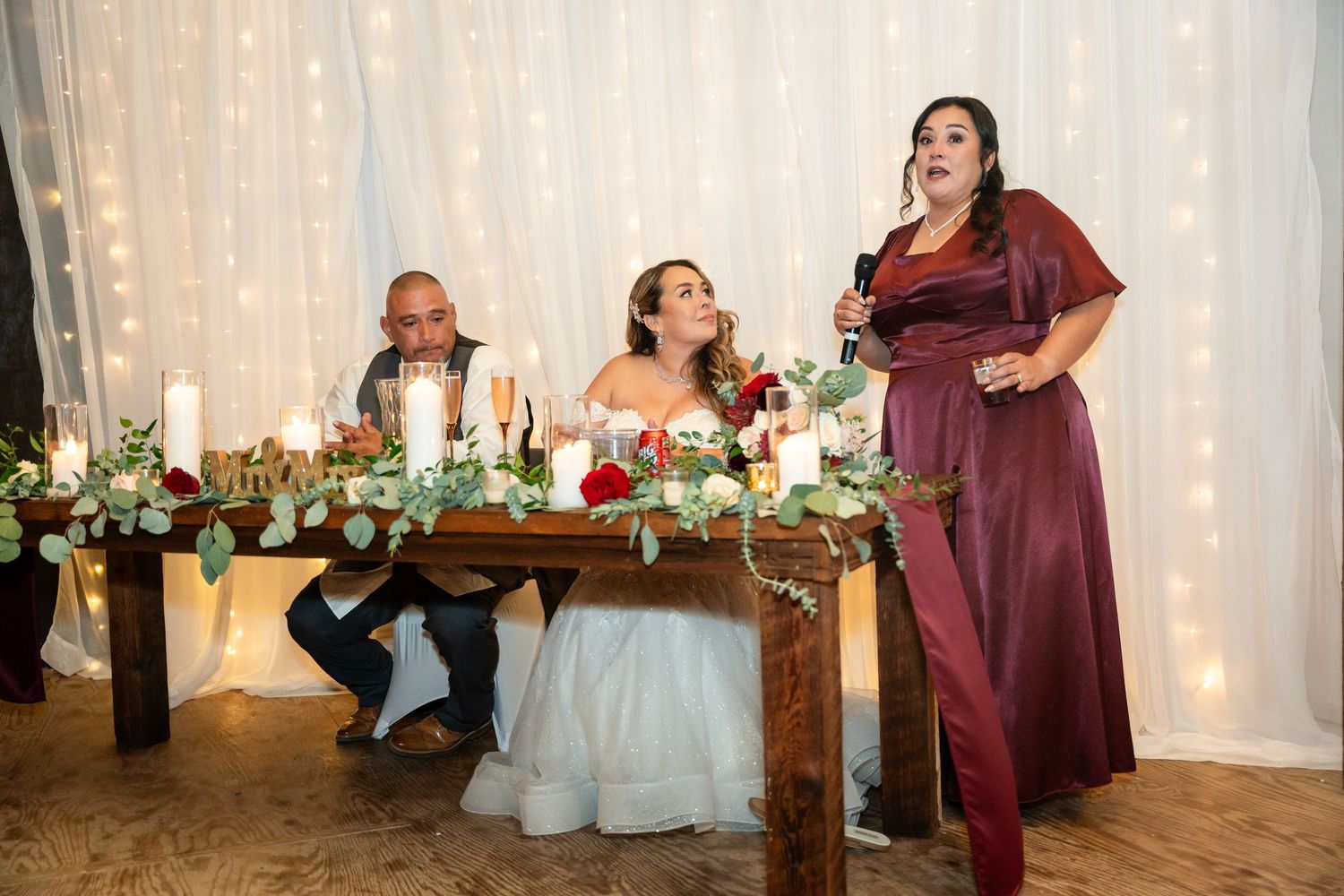 Wedding party members at the head table react during reception speeches against a backdrop of string lights.