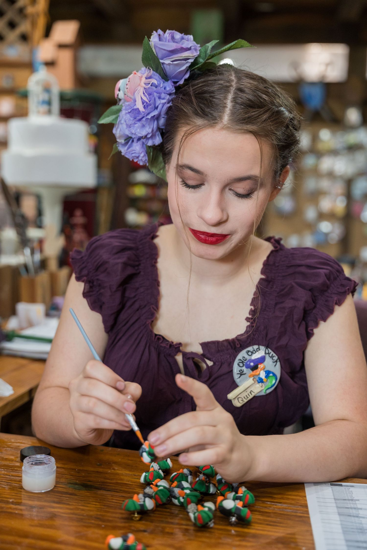 An artisan in a purple dress and floral hairpiece carefully paints detailed decorations at a wooden table.