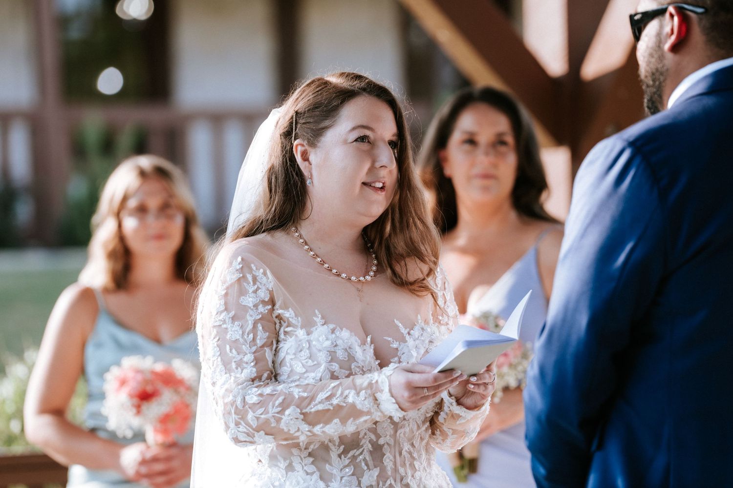 A bride in a lace wedding dress reads vows during an outdoor ceremony.