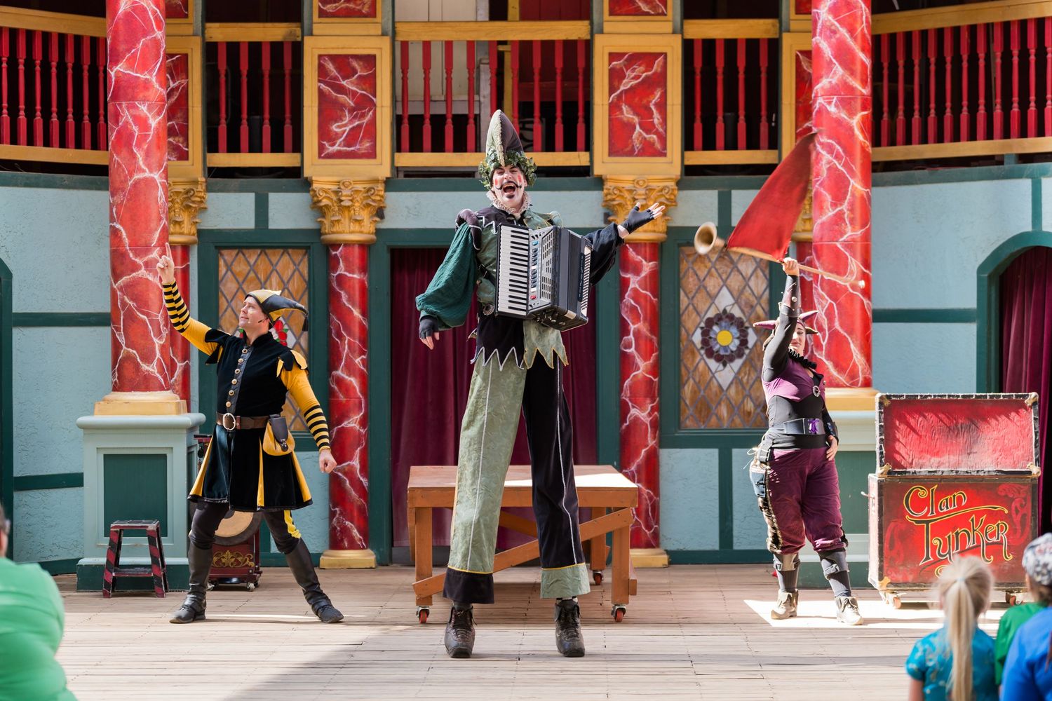 Three performers in colorful costumes dance and play accordion on a decorated stage.