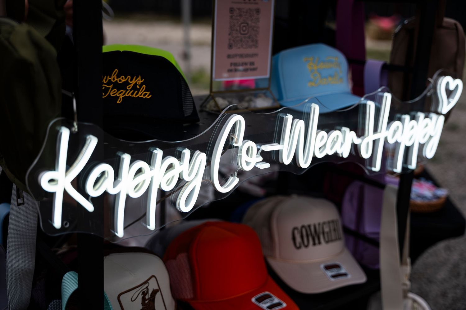 Neon sign reading 'Kappy Co-Wear Happy' displayed above baseball caps in a retail store window.