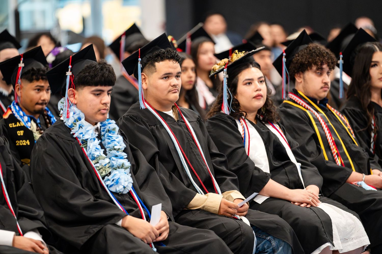 Graduates in black caps and gowns seated during commencement ceremony with decorative leis.