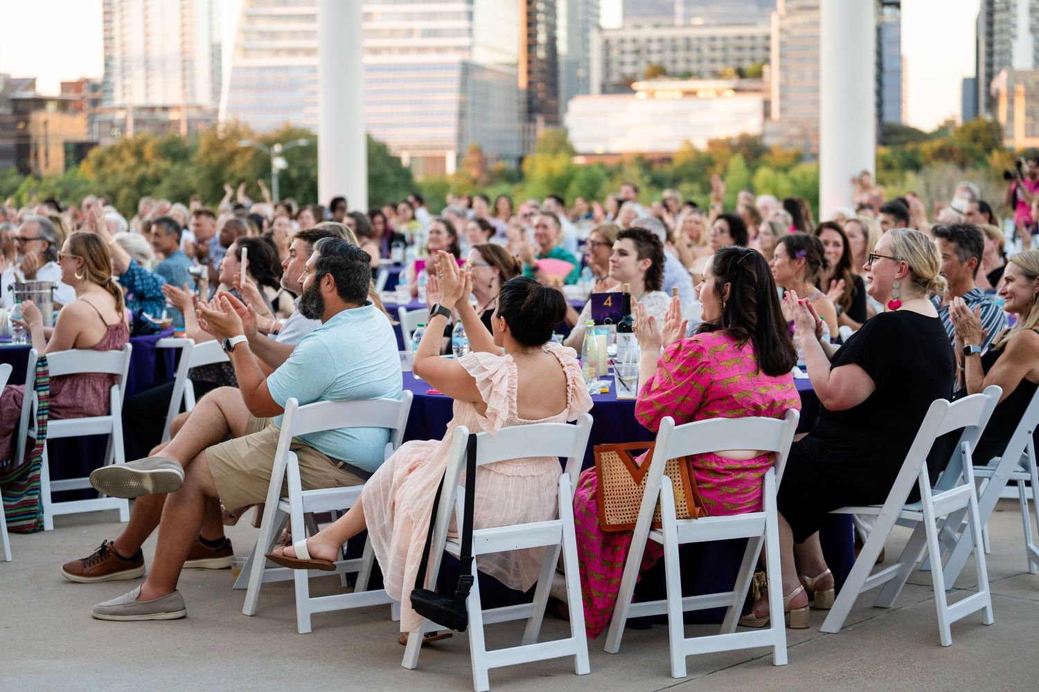 Diverse crowd seated in white chairs at outdoor event with city skyline in background.