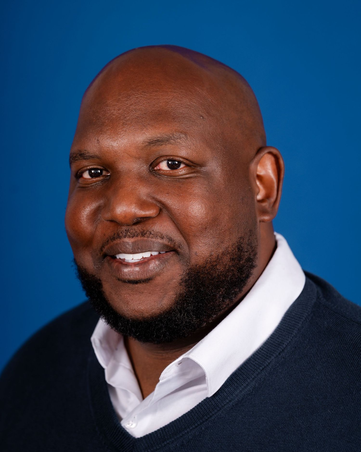 Professional headshot portrait of a smiling person wearing a black suit jacket with white shirt against a blue background.
