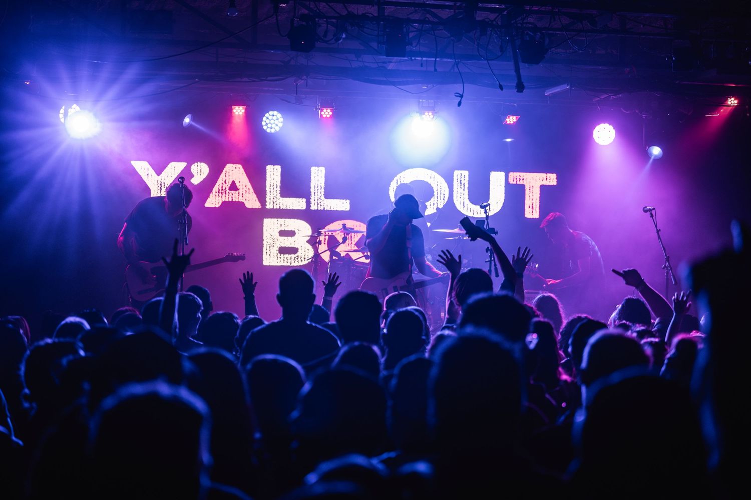 Concert crowd silhouettes dance under vibrant purple and blue stage lights with illuminated Y'ALL OUT text display.