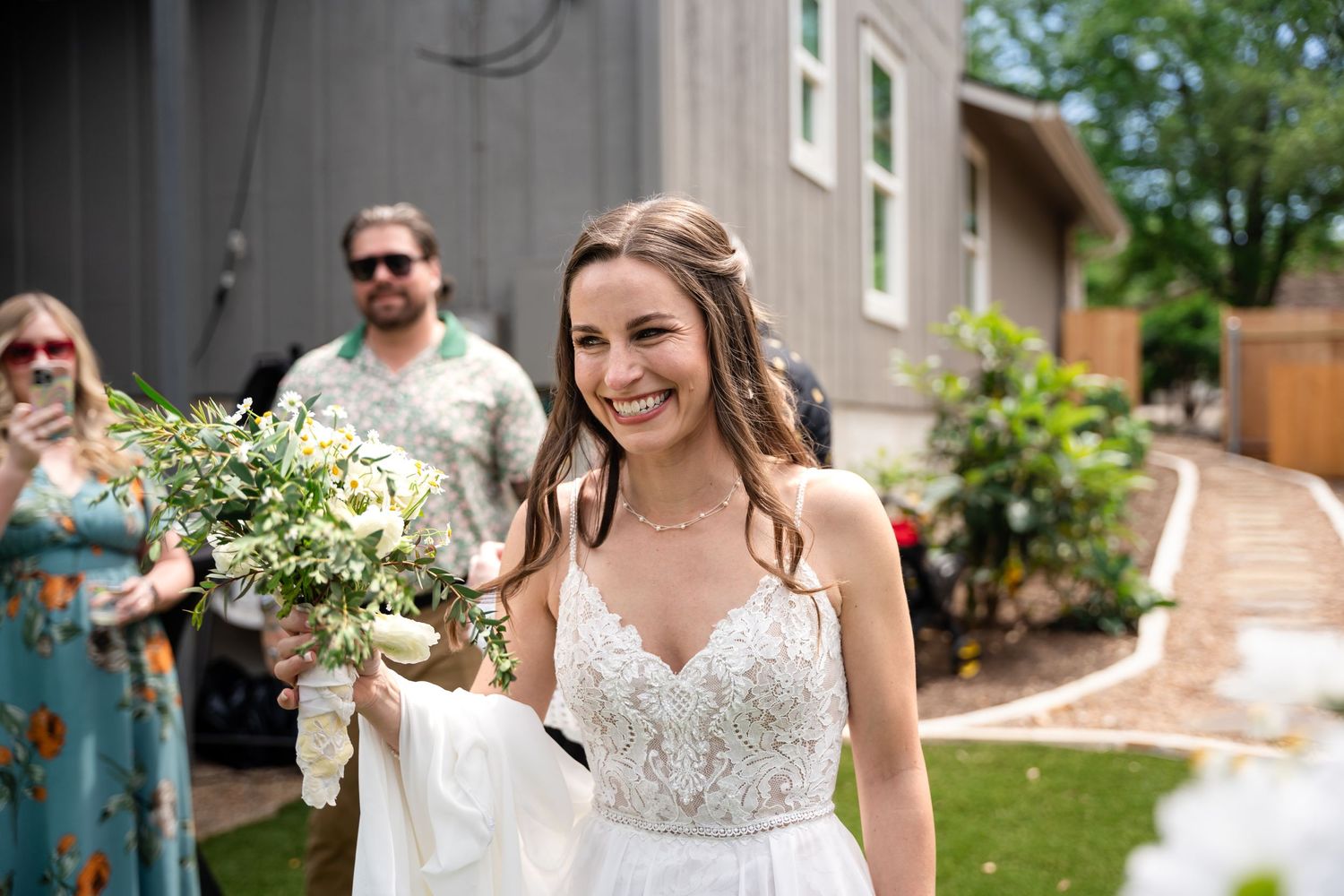 A bride holding a natural bouquet of white flowers smiles outdoors in a lace wedding dress.
