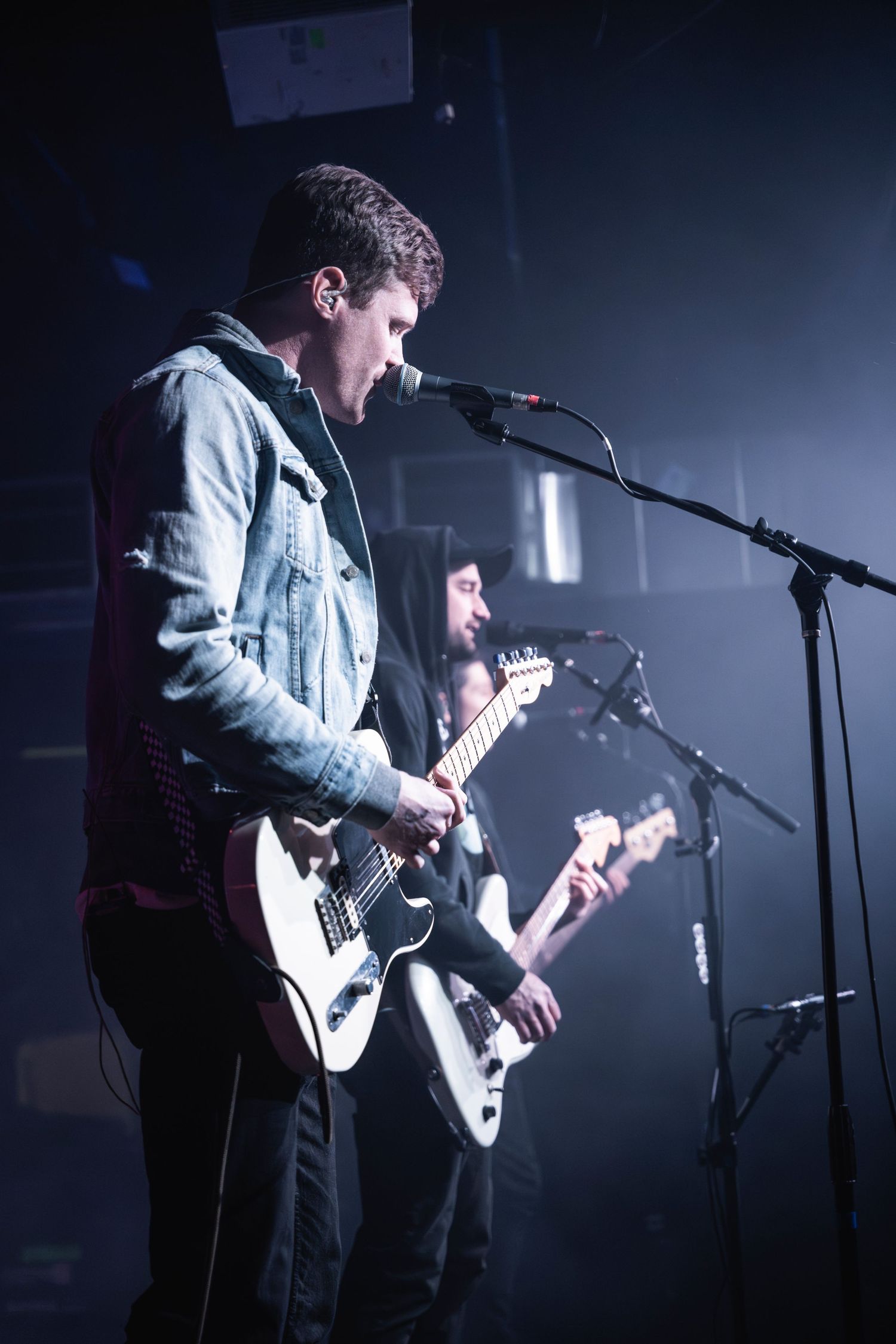 Guitarist in denim jacket performing with band under dramatic stage lighting.