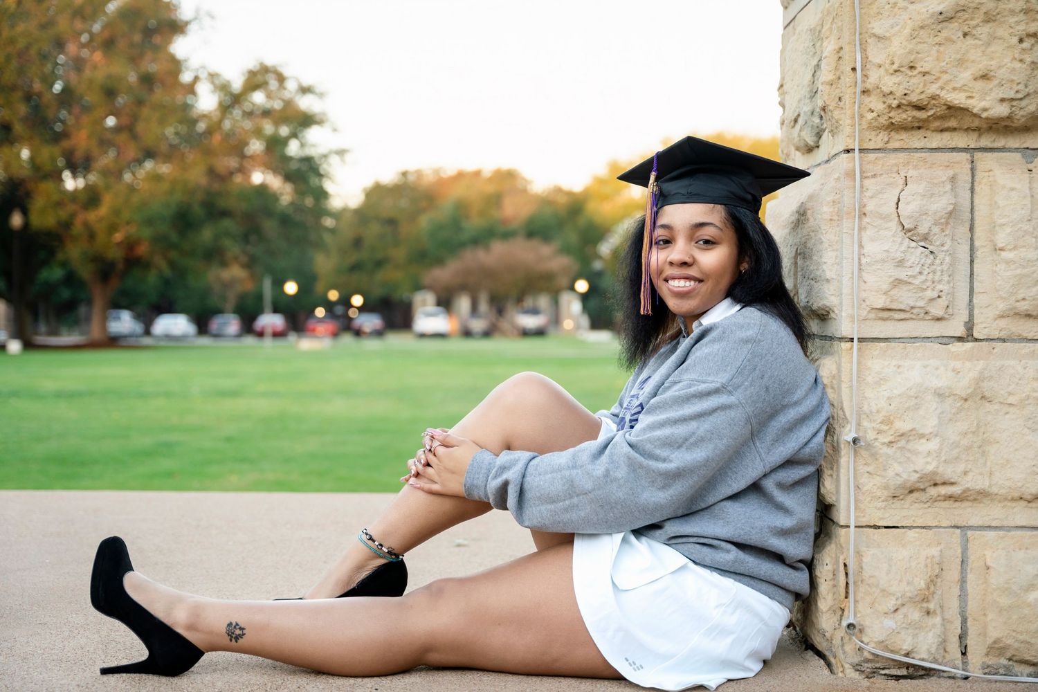 Graduate in cap and sweatshirt sits casually against stone wall during outdoor photoshoot.
