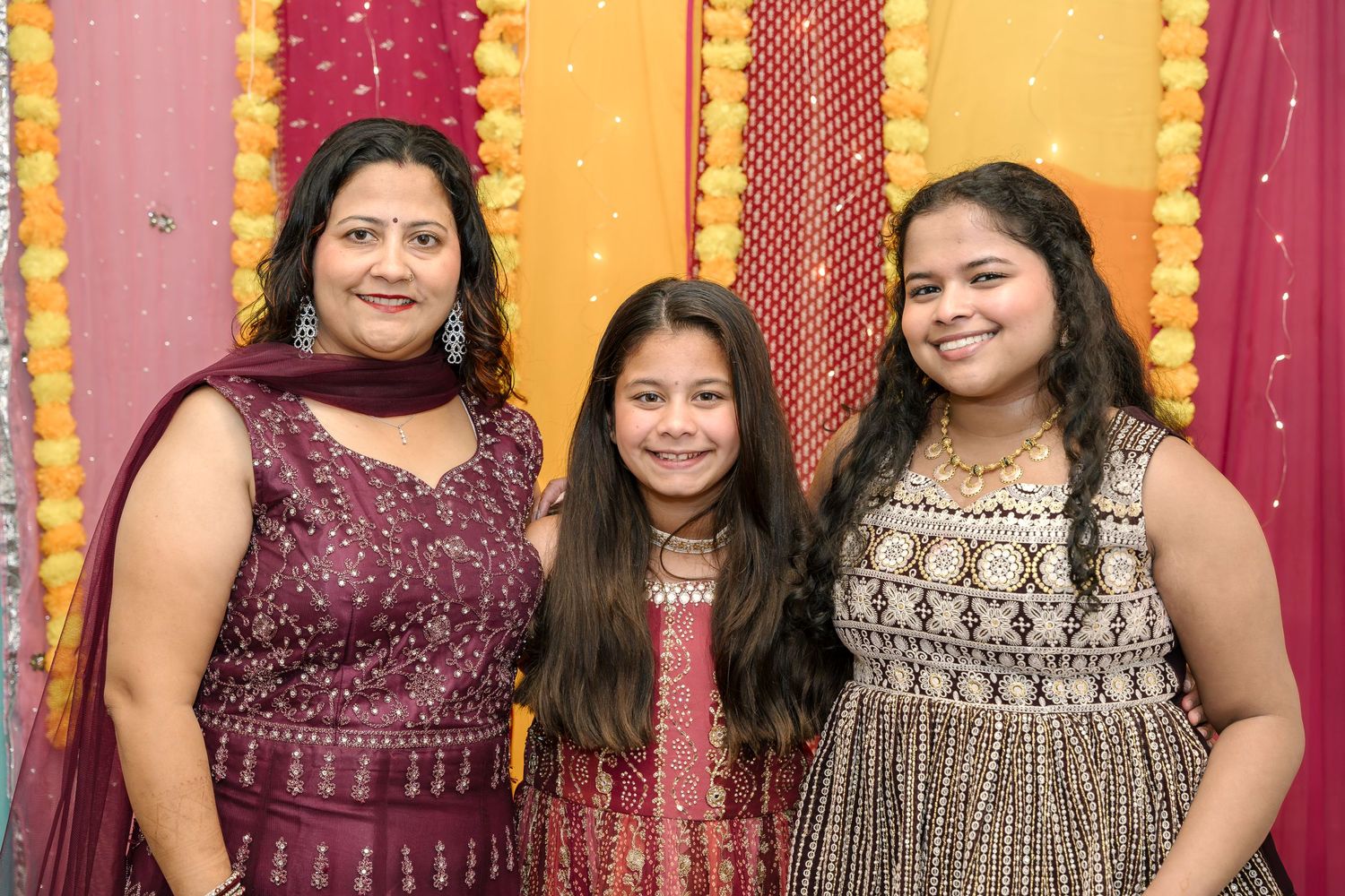 Three people in festive attire posing against decorative backdrop with orange and red draping.