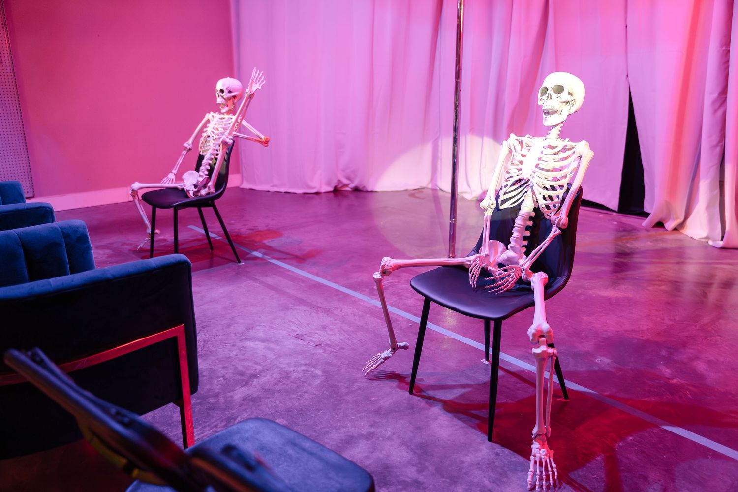 Plastic skeletons sit posed on chairs in a pink-lit theatrical scene.