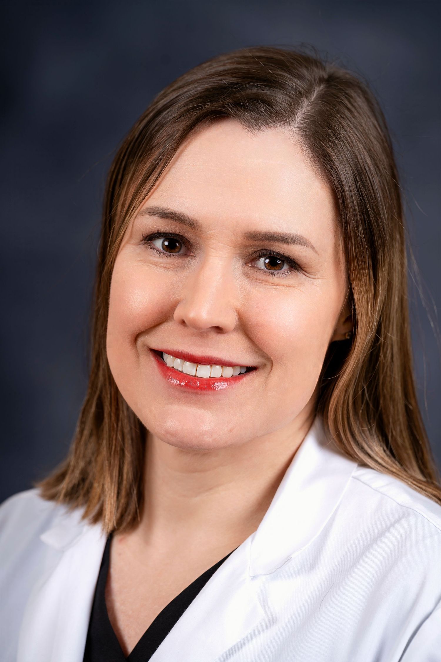 Professional portrait of a medical worker wearing a white coat and dark top against muted background.
