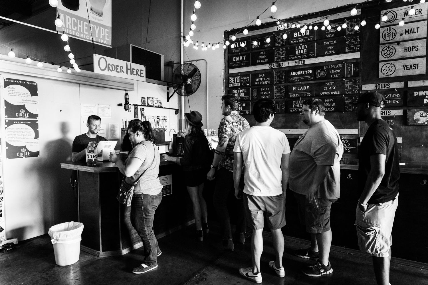 Customers wait in line at a bar counter with chalkboard menus and string lights overhead.
