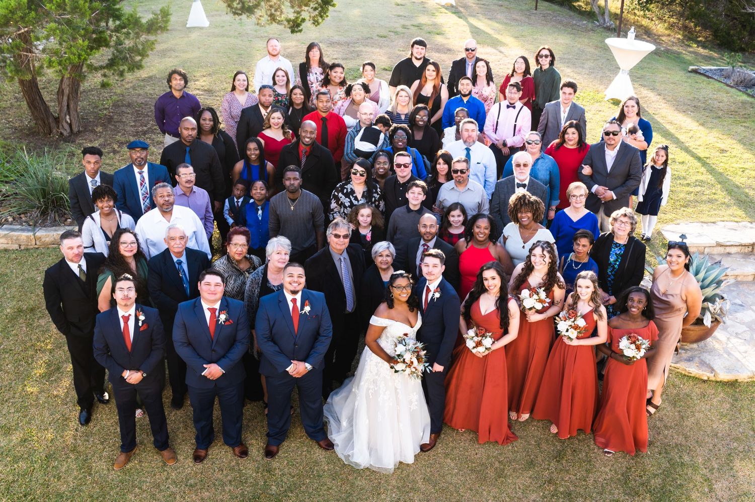 Large wedding party and guests group photo taken outdoors with bridesmaids in rust-colored dresses.