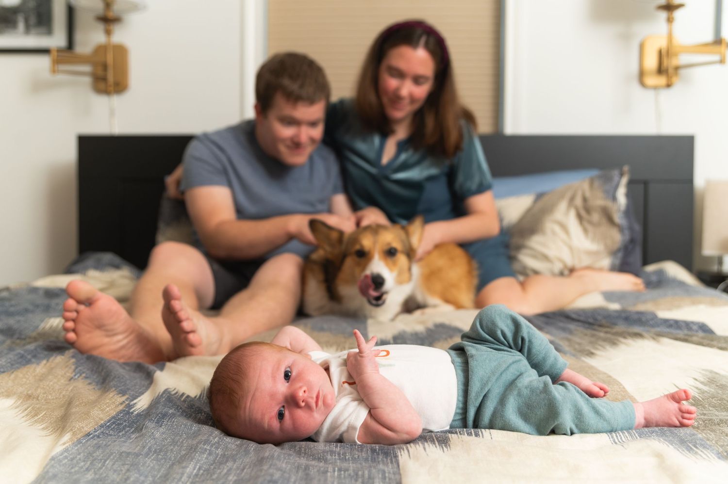 Family relaxes on bed with dog while baby lies on blanket.