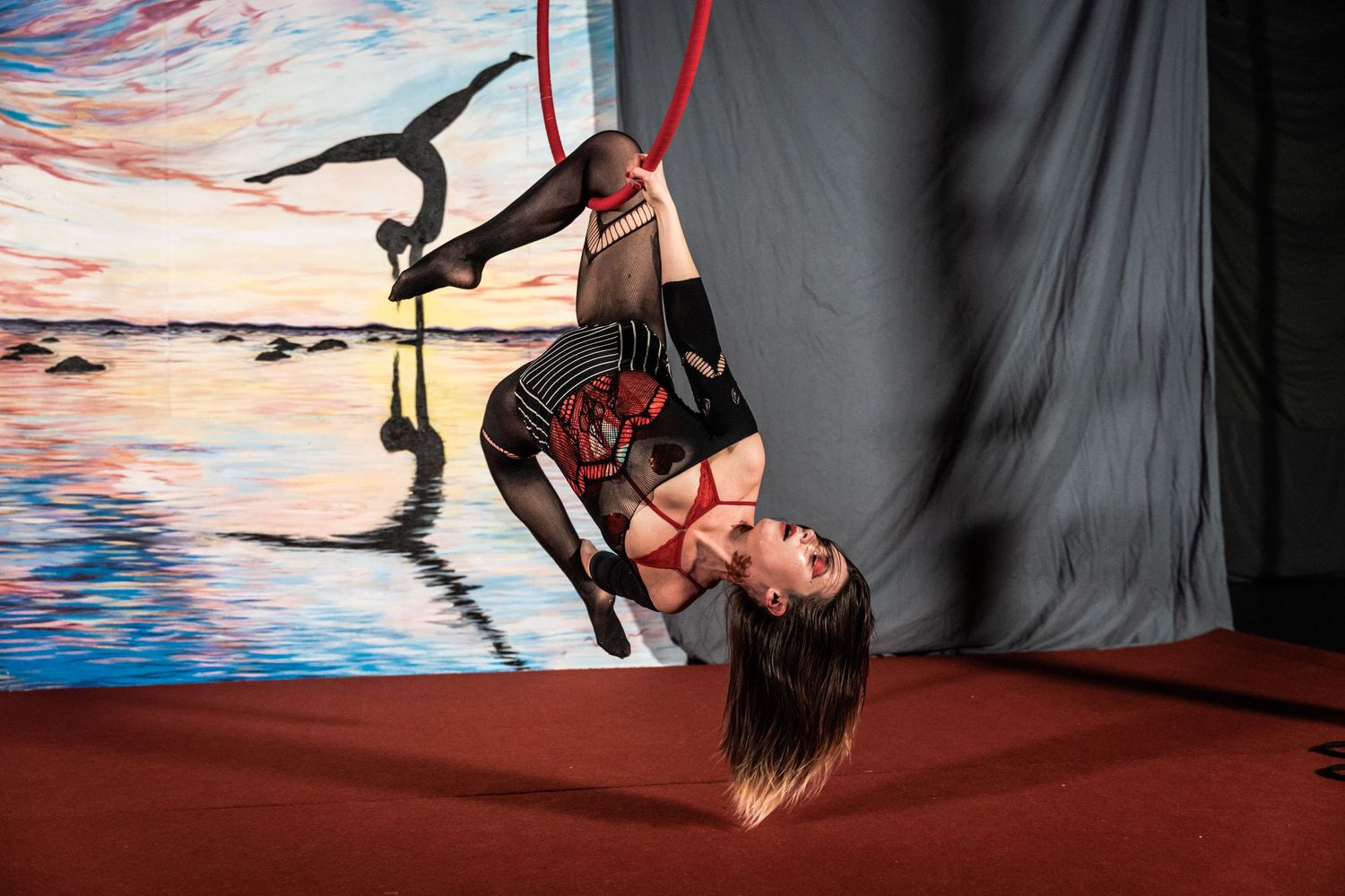 Aerial silk performer executing an inverted pose against painted sunset backdrop.