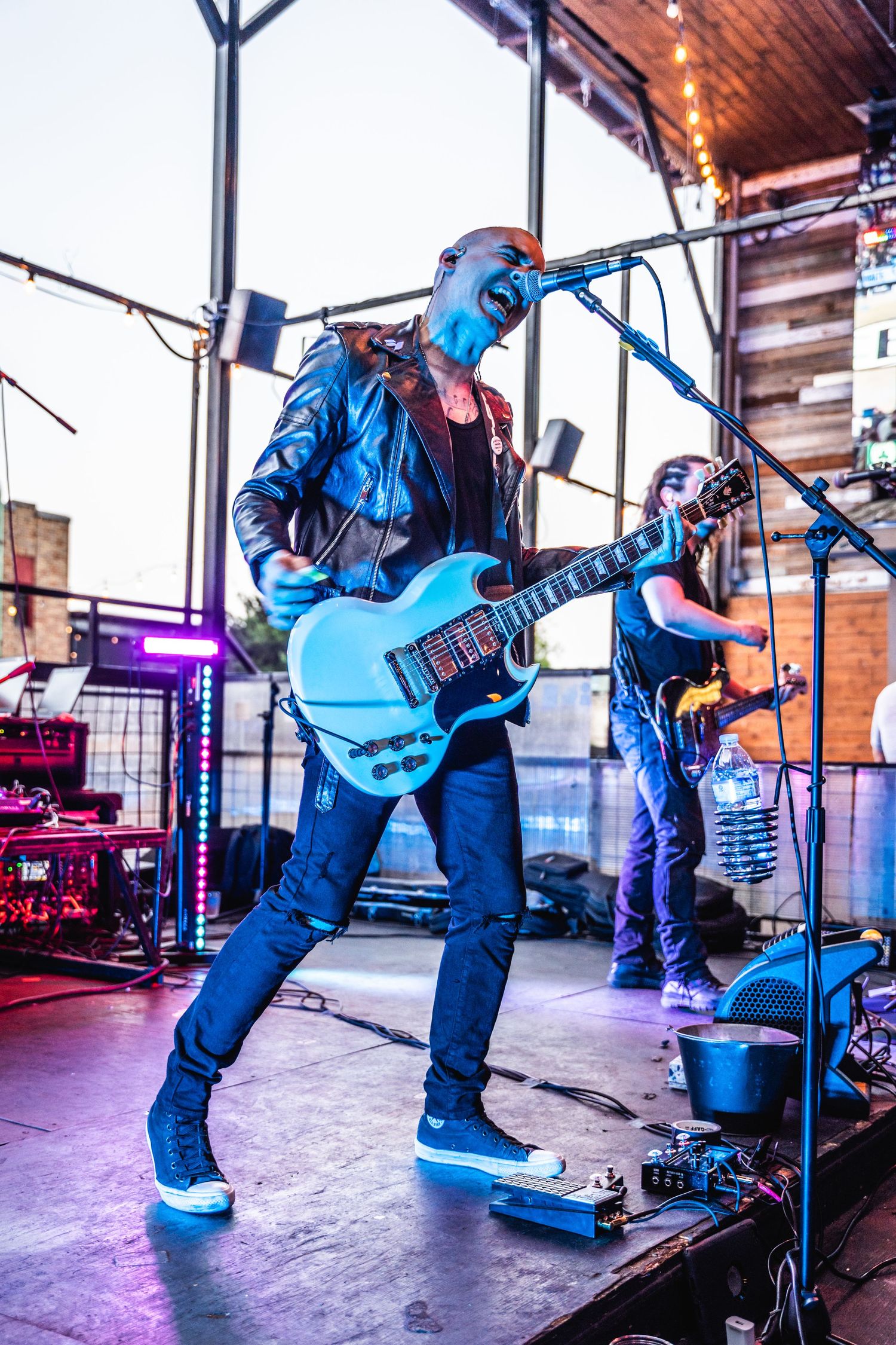 Electric guitarist performs energetically on stage in blue lighting with band members visible in background.