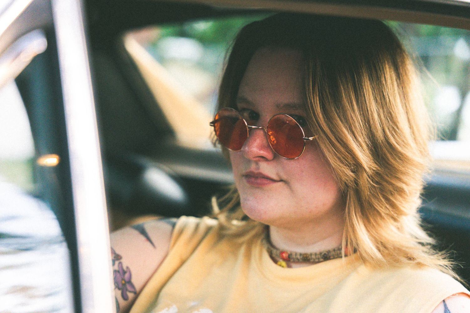 Person in car wearing retro sunglasses and yellow shirt with sunlight streaming through window.