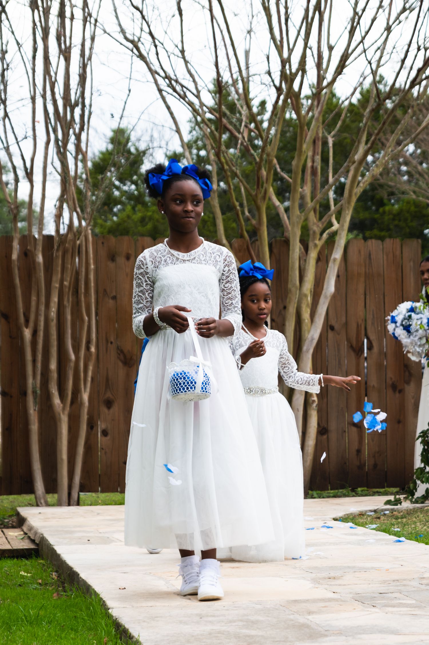 Two flower girls in white dresses and blue hair accessories walk down an outdoor path carrying small bouquets.