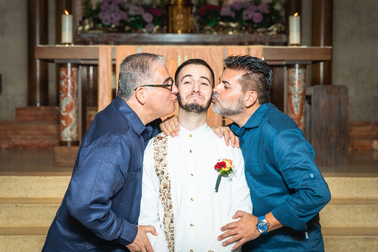 A groom receives affectionate kisses on the cheeks from two others at a church altar.