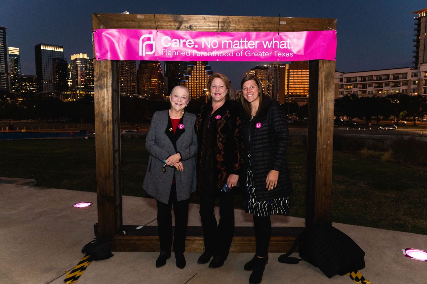 Three people stand under pink illuminated sign at nighttime outdoor event.