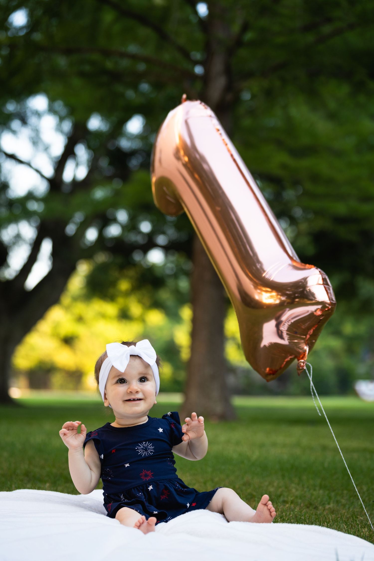 Baby in navy dress sits on blanket with rose gold shoe-shaped balloon in park setting.