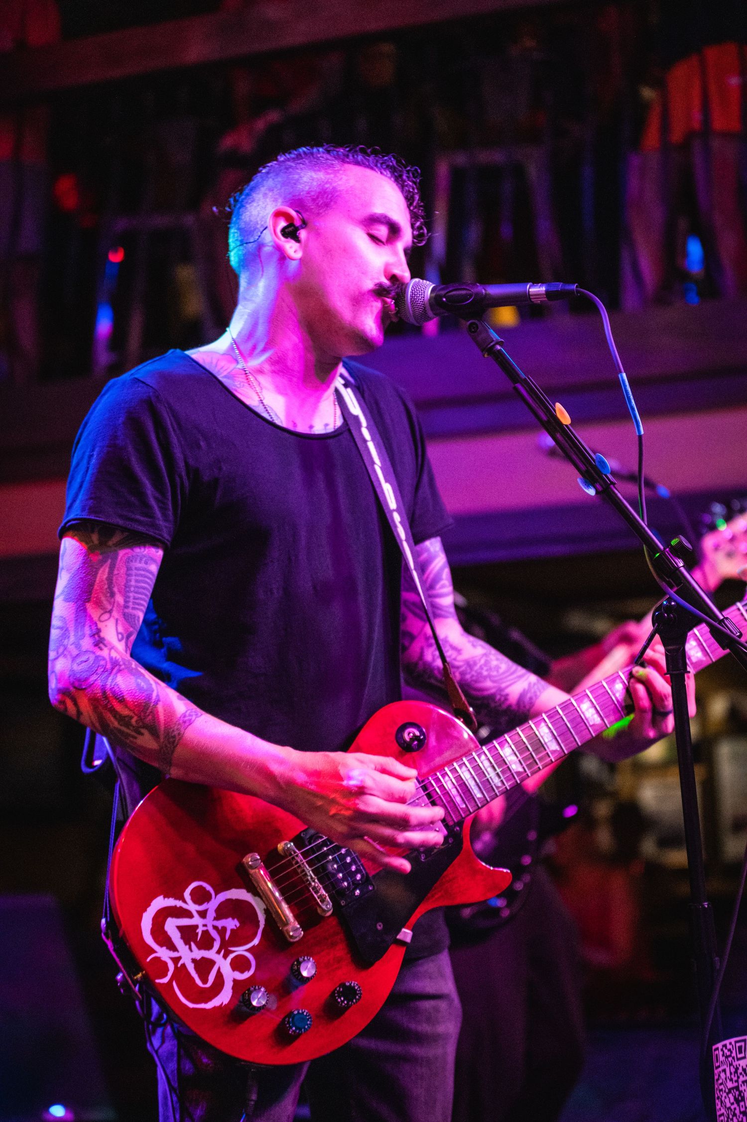 Musician in black t-shirt plays red guitar with decorative pattern under purple stage lights.