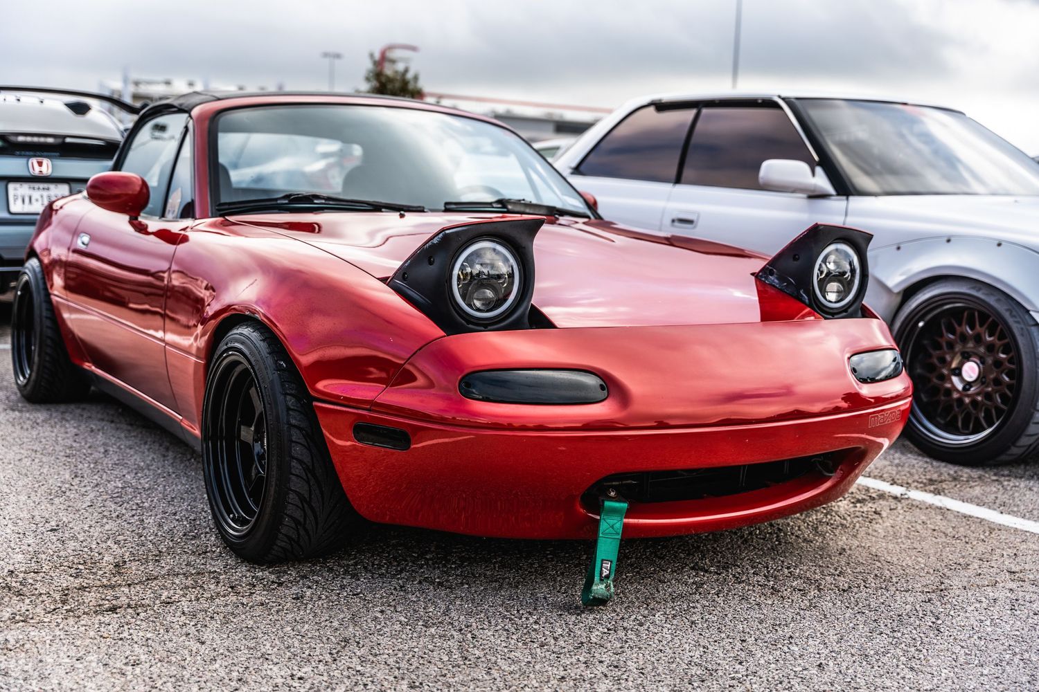 Red Mazda Miata with pop-up headlights and black wheels parked at a car show.