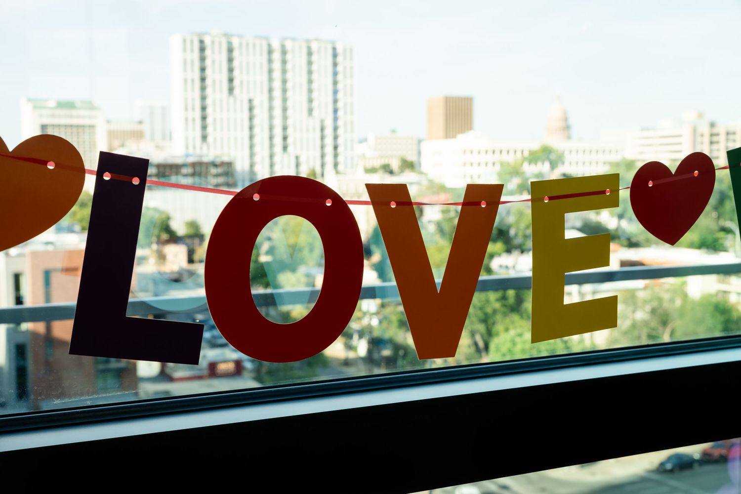Colorful LOVE letter banner with heart decorations hanging on a window with city skyline view.