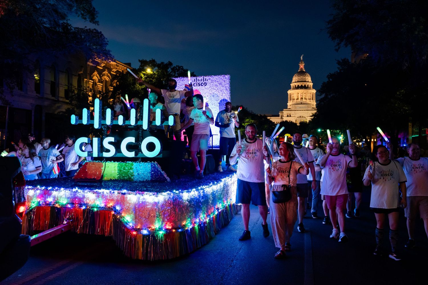Cisco illuminated float with colorful lights leads a night parade near a capitol building.