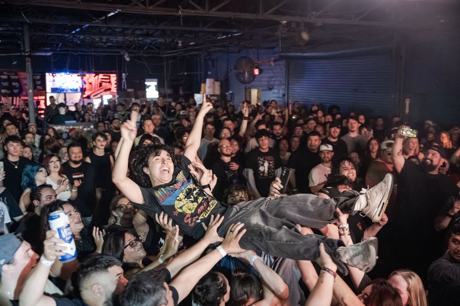 Crowd surfing at a packed indoor metal concert with fans in black shirts cheering under bright stage lights.