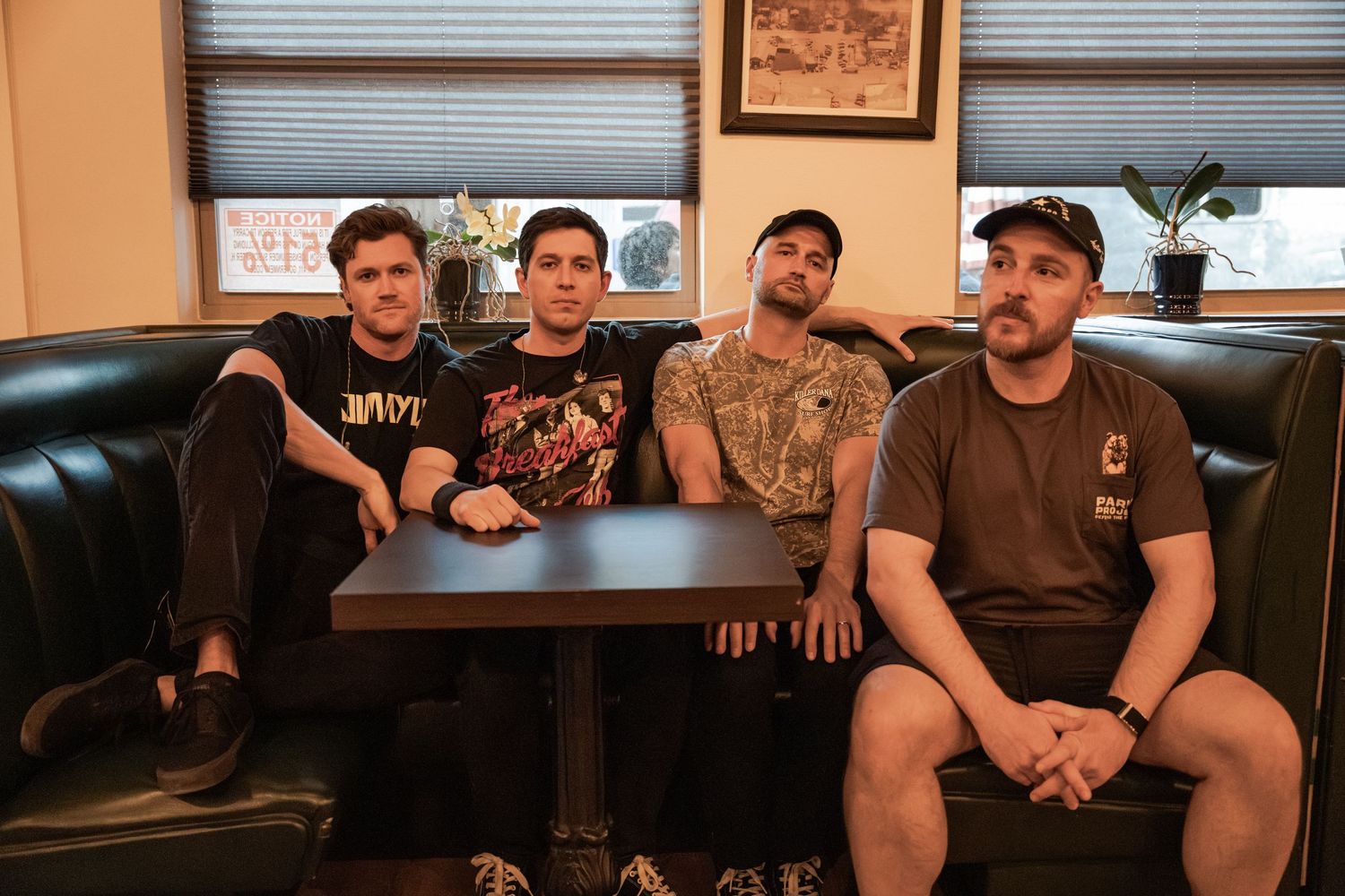 Four young men sitting together in a diner booth, wearing casual band t-shirts, posing for a group photo.