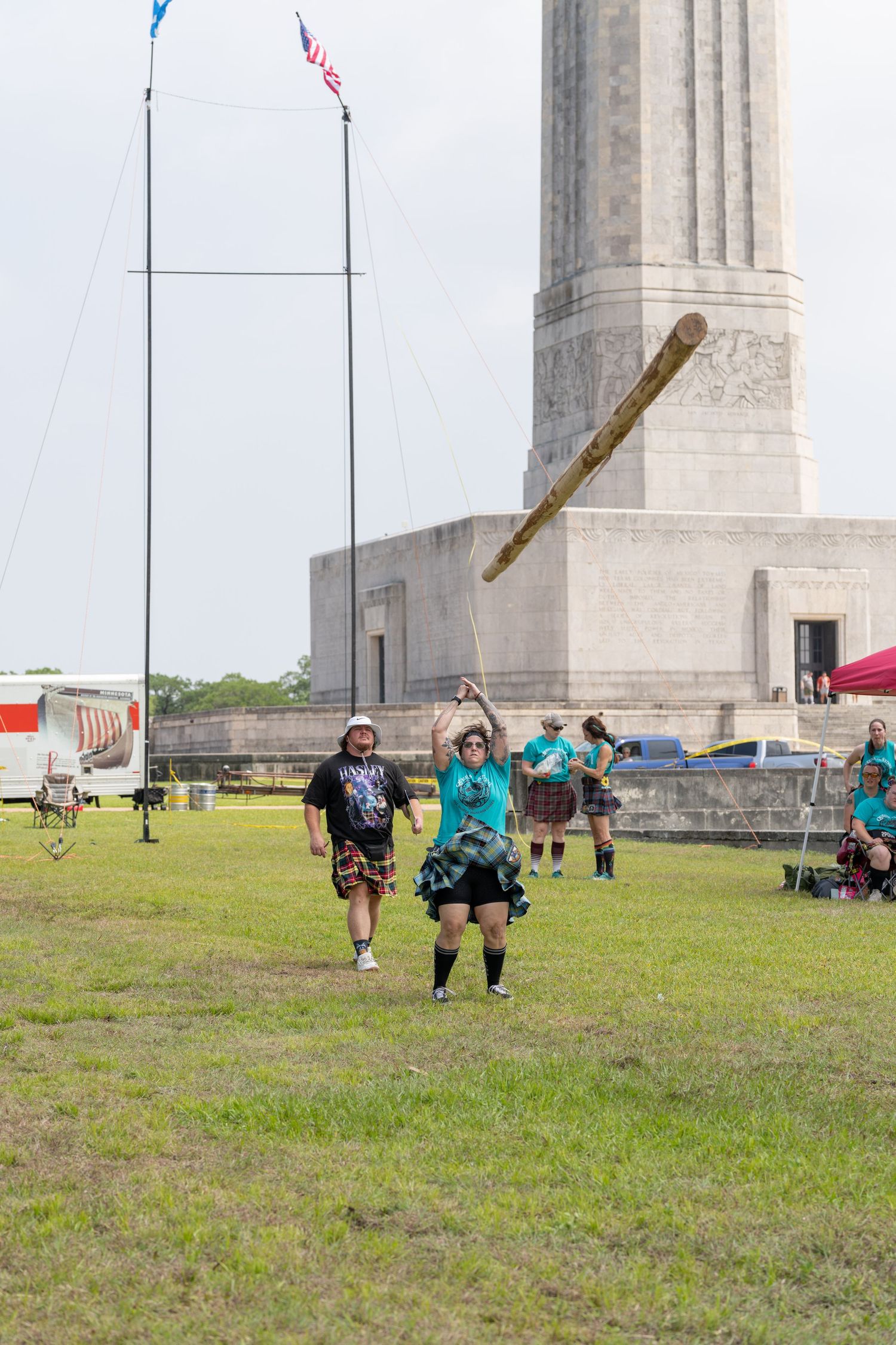 Woman tosses a caber at a Highland Games competition near a stone monument with spectators watching.