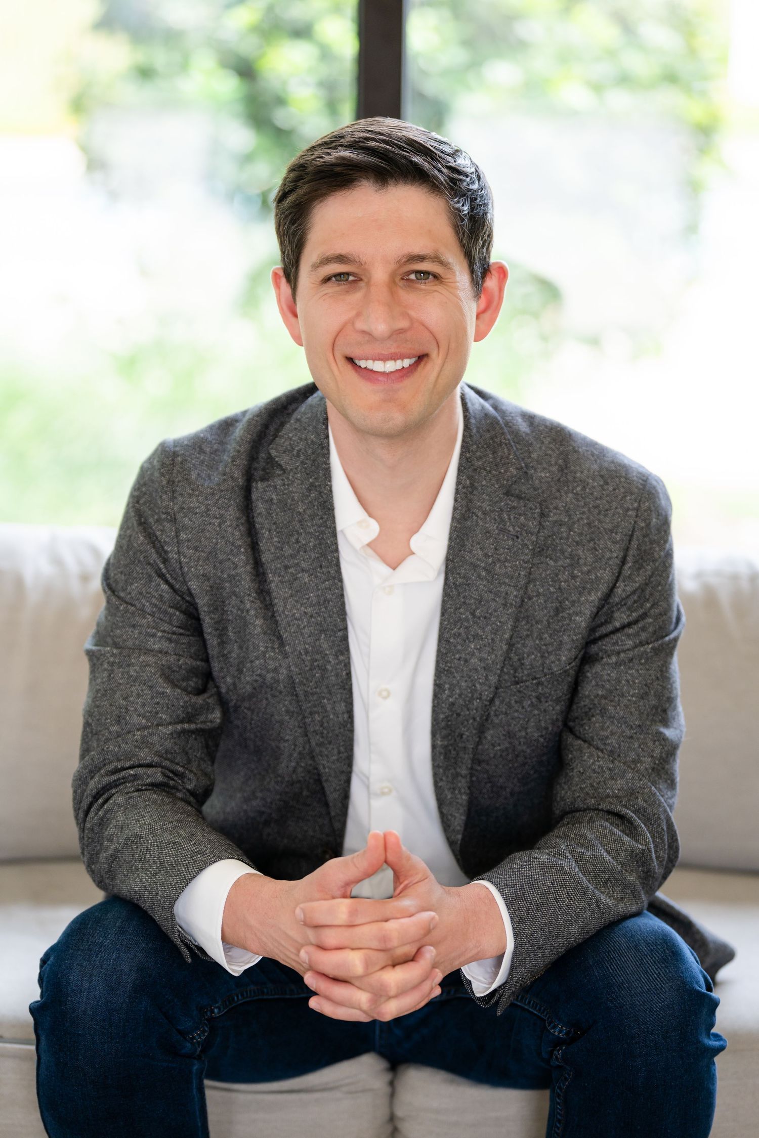 Smiling man in gray blazer and white shirt sits on sofa with hands clasped, bright window background.