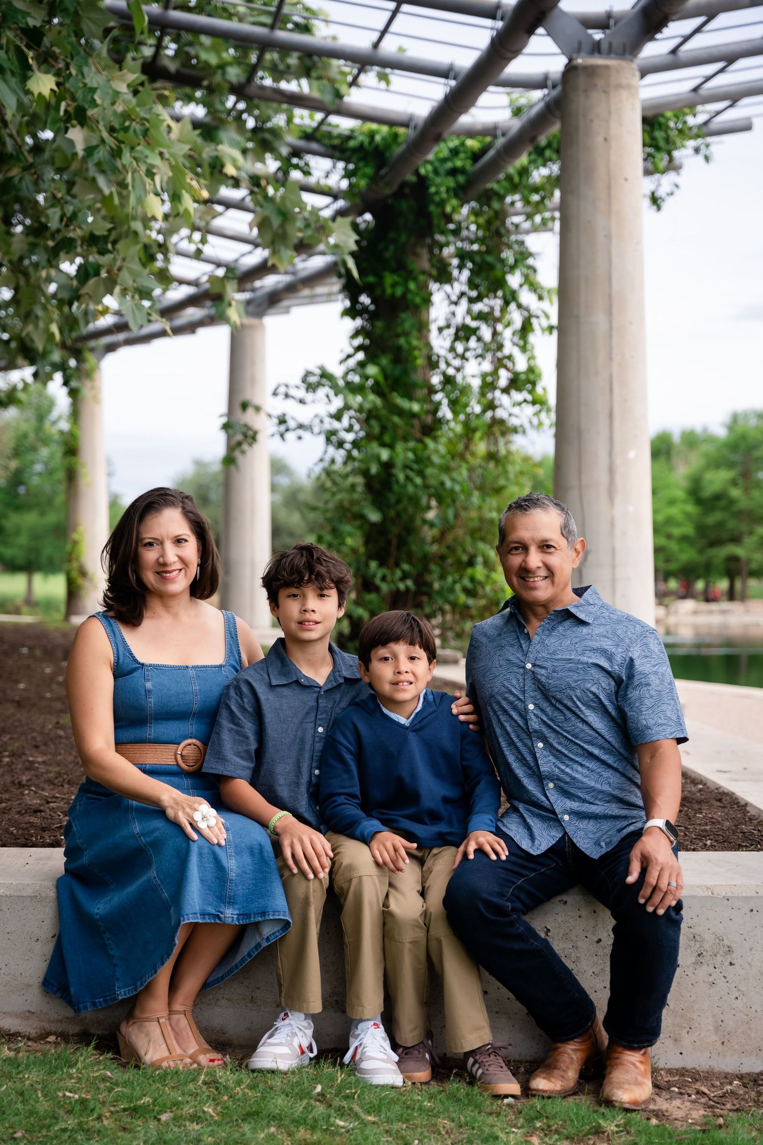 Family of four smiling together under a pergola in a lush green park, wearing coordinated blue and tan outfits.