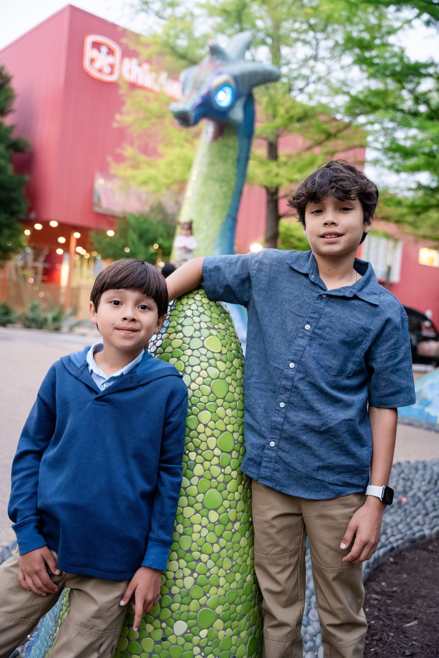 Two boys posing with a colorful green dinosaur sculpture outside a red building surrounded by trees.
