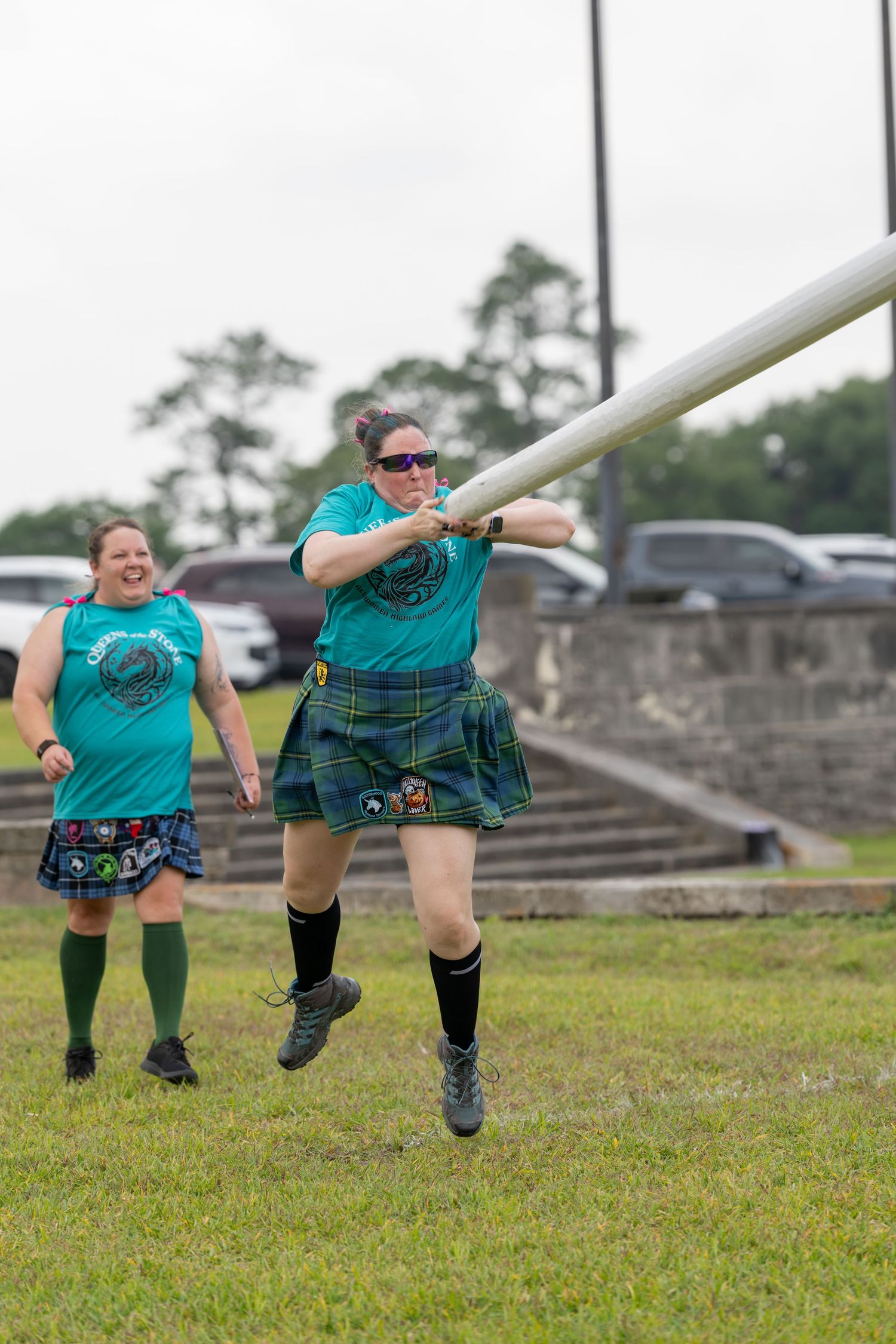 Woman in tartan kilt tosses caber at Highland Games competition while another competitor watches and laughs.