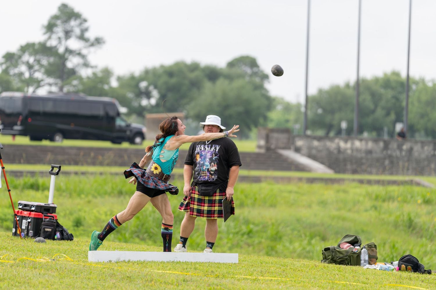 Woman throws shot put at Highland Games while man in kilt watches on green field.