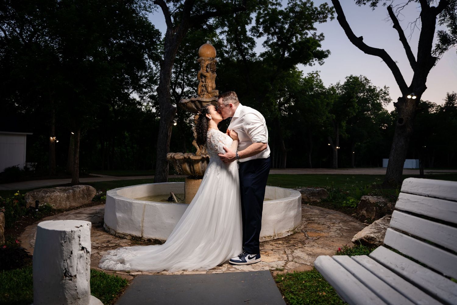 Bride and groom share a romantic kiss near a stone fountain at dusk in a lush park setting.