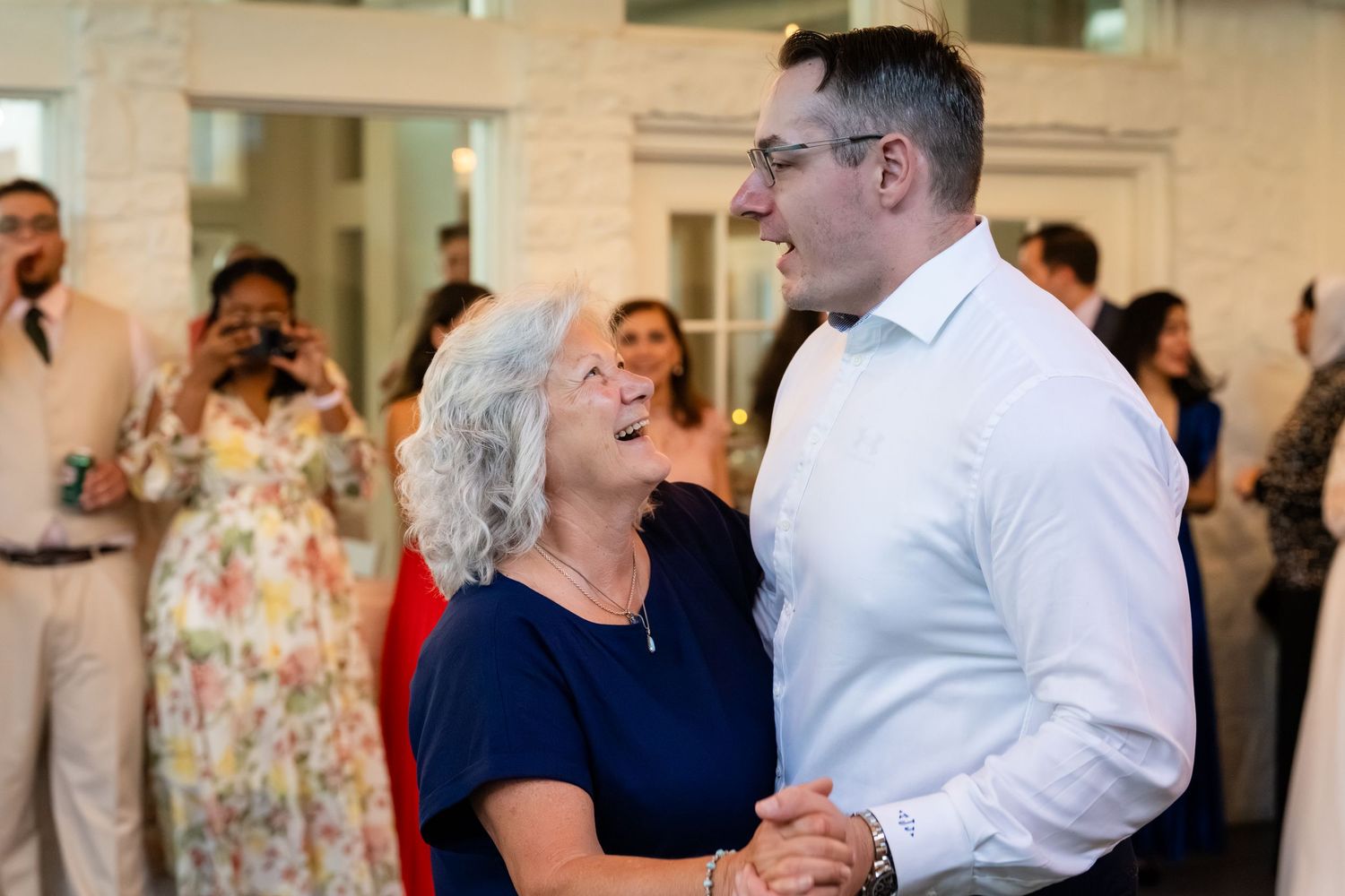 A joyful mother-son dance at a wedding reception, with guests photographing the heartwarming moment in an elegant venue.